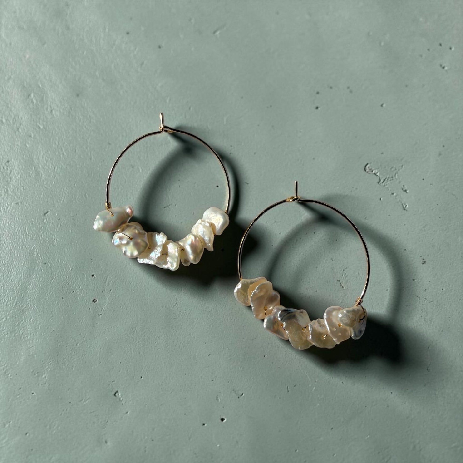 TNT Designs Jewels gold hoop earrings featuring lustrous irregular baroque pearls on a sage green textured background.